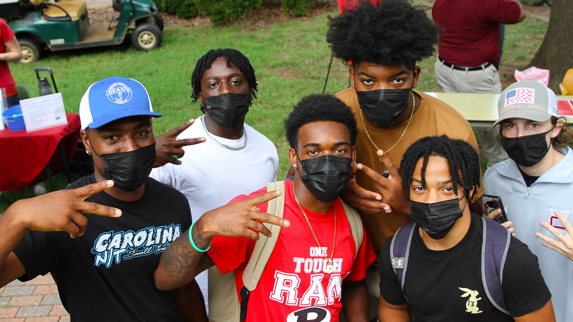 Five students wearing masks stand together to take a photo, showing peace signs with their fingers.