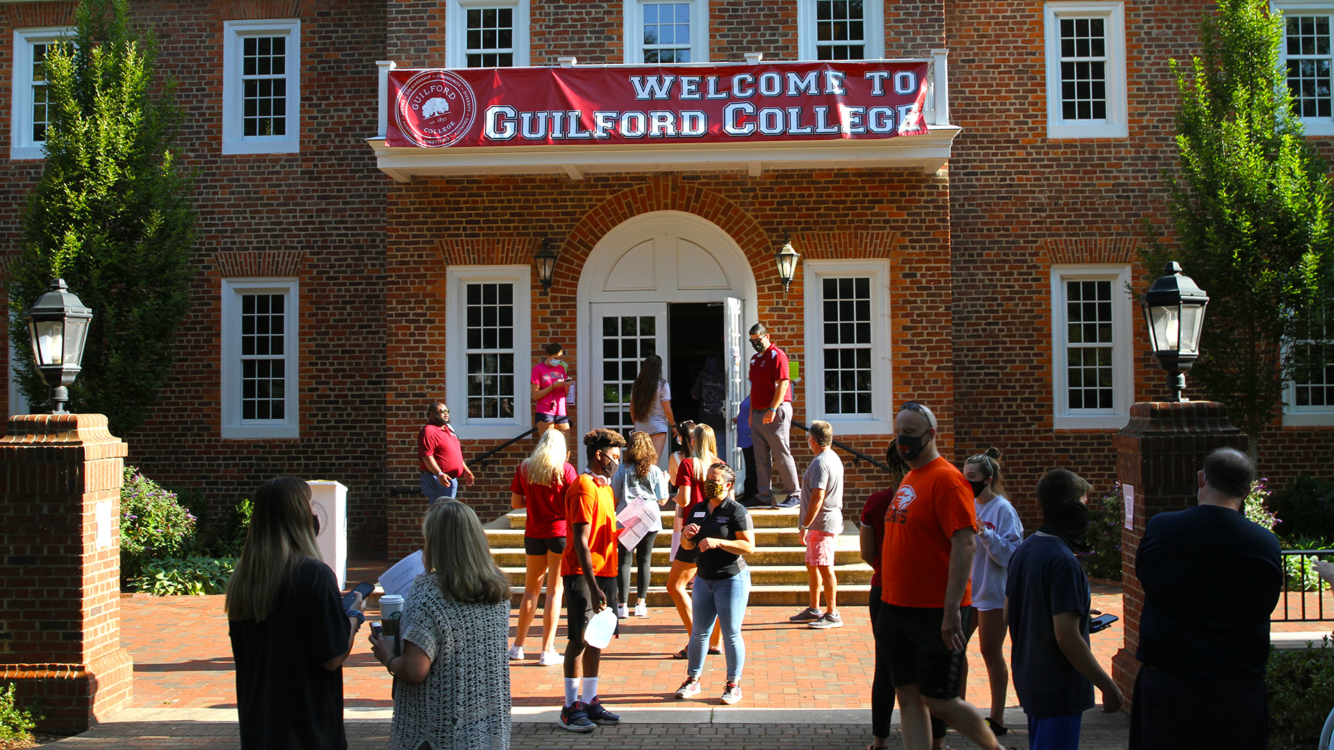 Students and families arrive at Founders Hall on move-in day, where a banner saying Welcome to Guilford College hangs on the balcony.