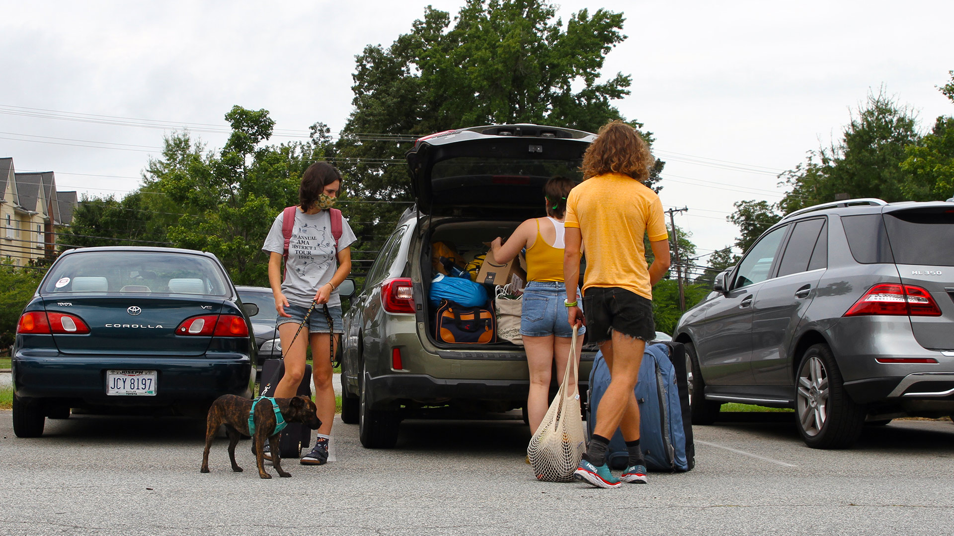 Three students, one holding a dog on a leash, unpack belongings from the back of a car.