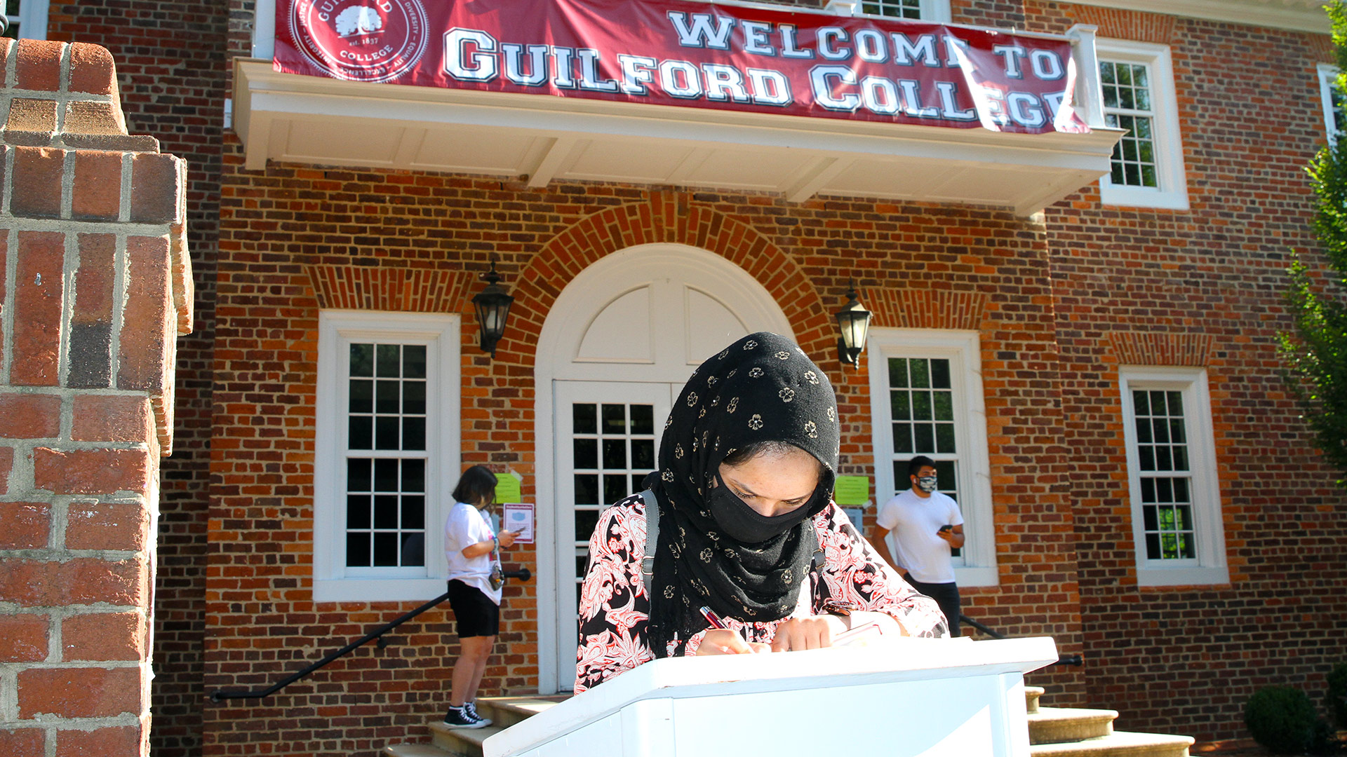 A student wearing a black head wrap decorated with tiny white flowers signs the registration book.