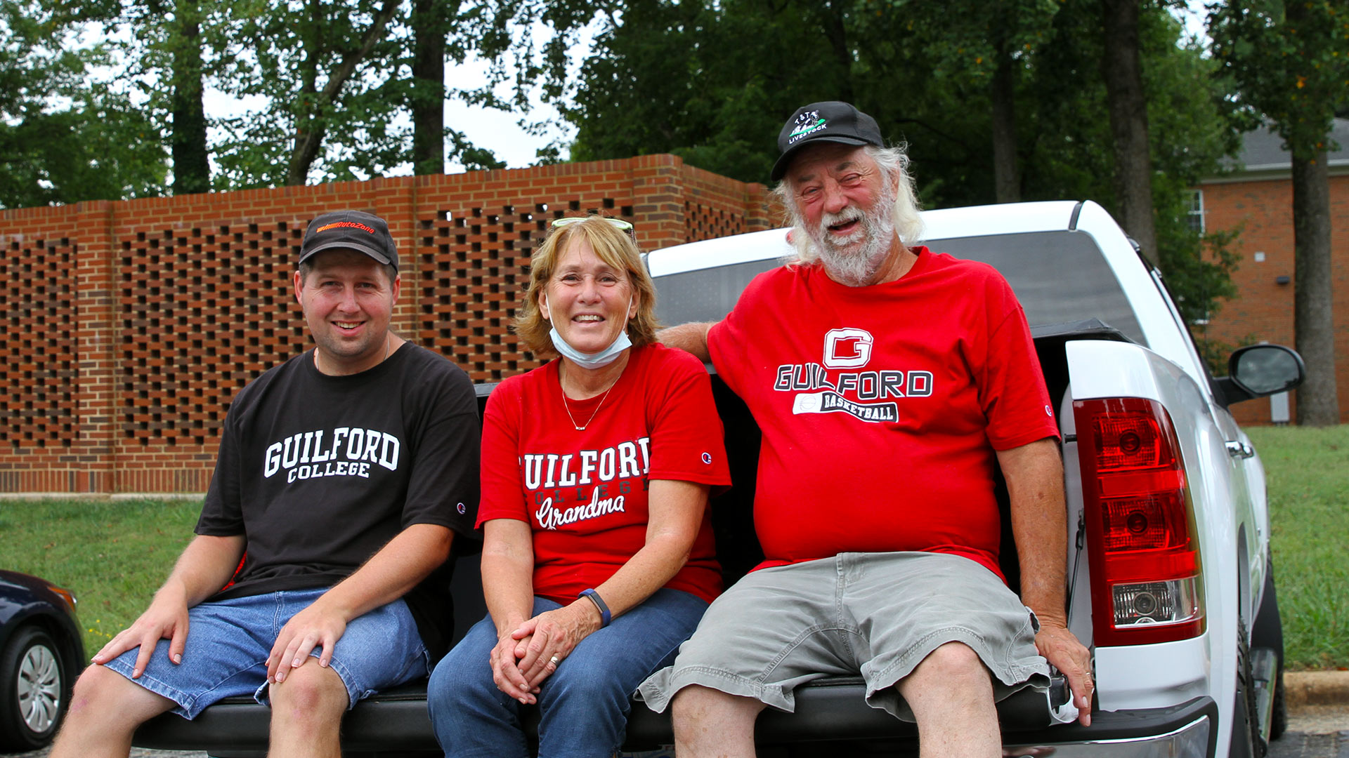 A family of three all wearing Guilford t-shirts smiles at the camera as they sit on a truck tailgate after moving in a student.