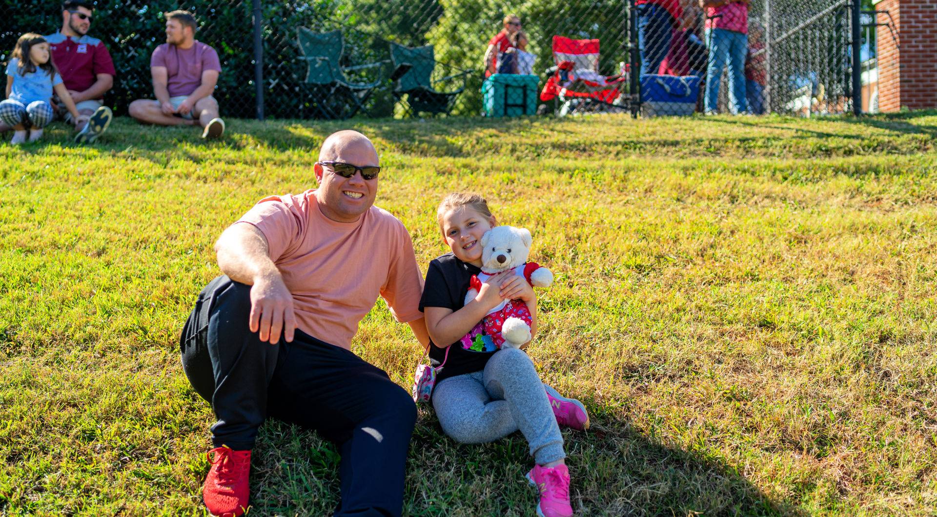 A parent and child, holding a teddy bear, watch the game from the hill.