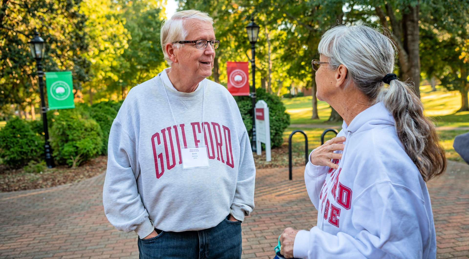 Two Guilfordians wearing gray Guilford College sweatshirts chat on the Quad.