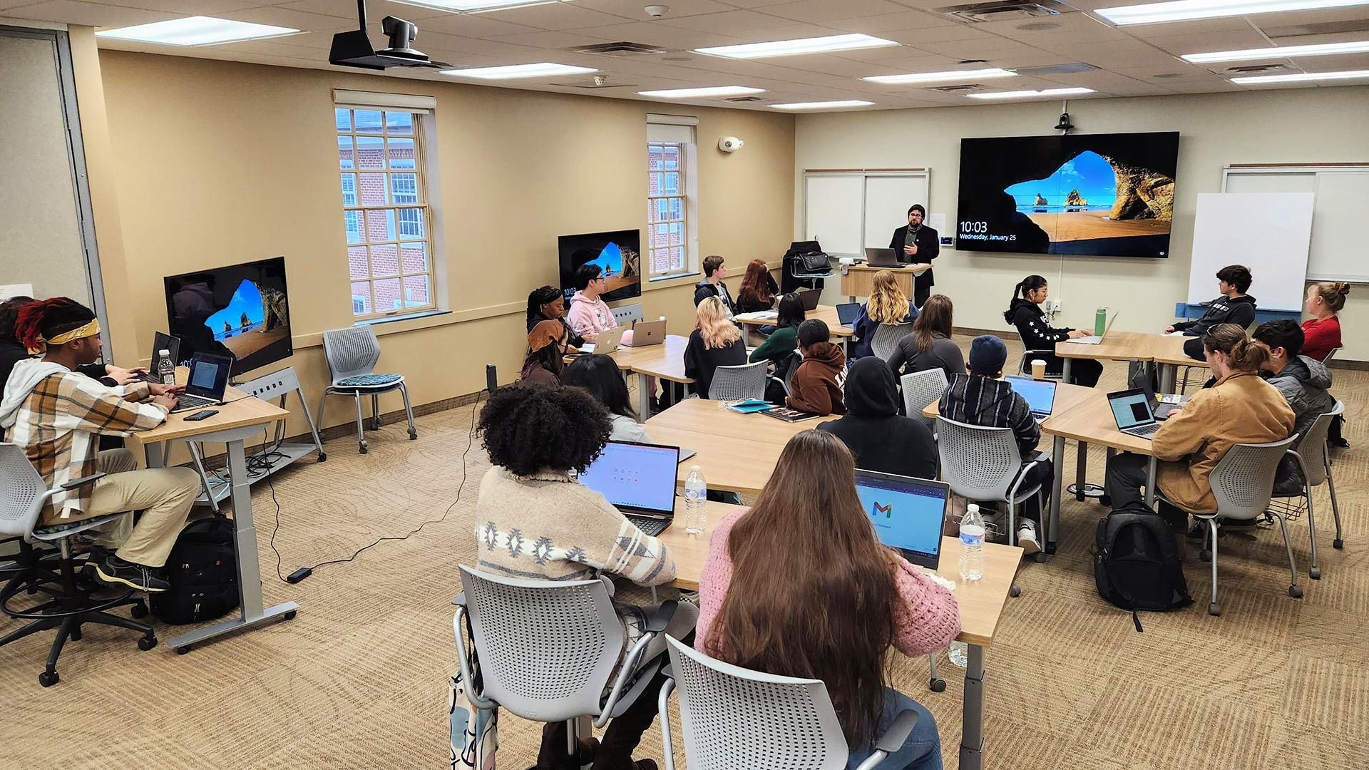 A professor stands at a podium in the Collaboratory, while students fill the seats, organized in pairs and small groups, in moveable chairs around the room.