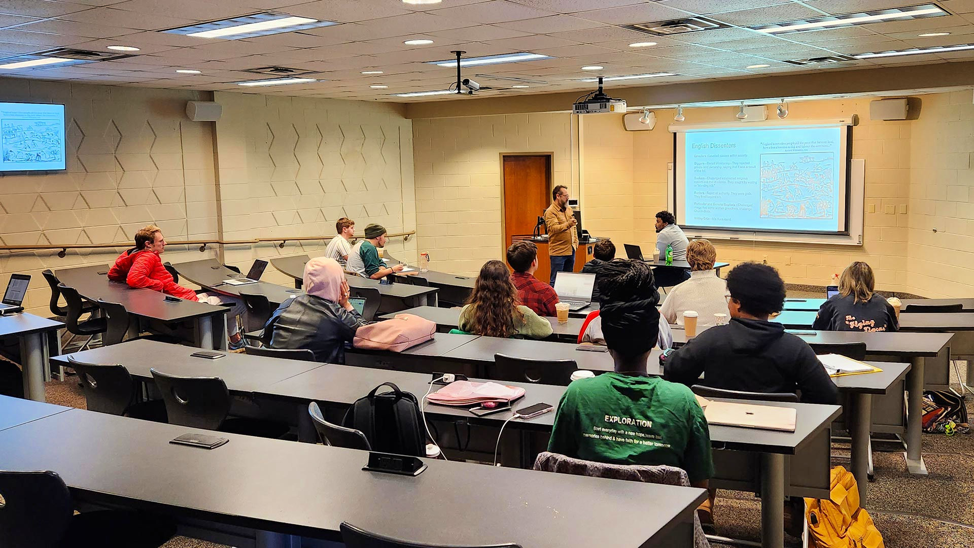 A professor stands at the front of King Hall 123 in front of a projector screen, while students fill the seats on graduated rows.