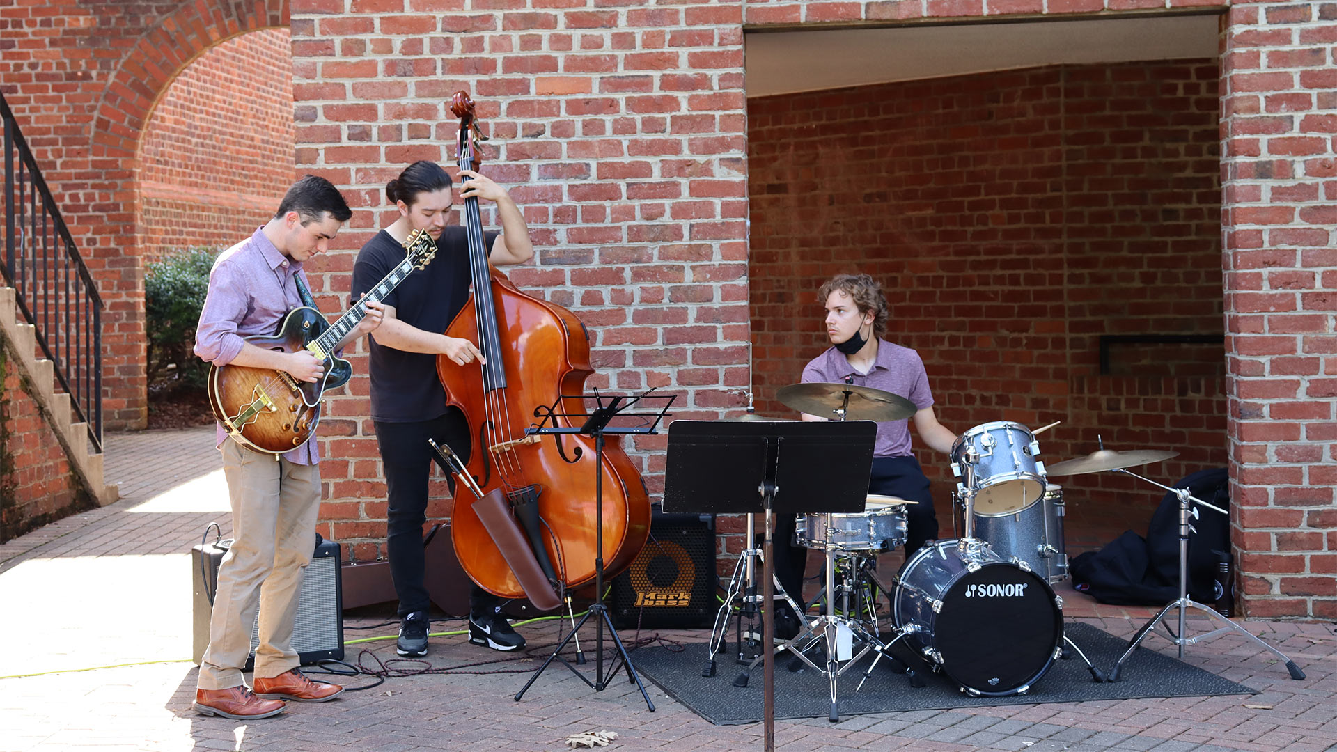 Student musicians play the guitar, standing bass, and drums on the patio beside Founders Hall.